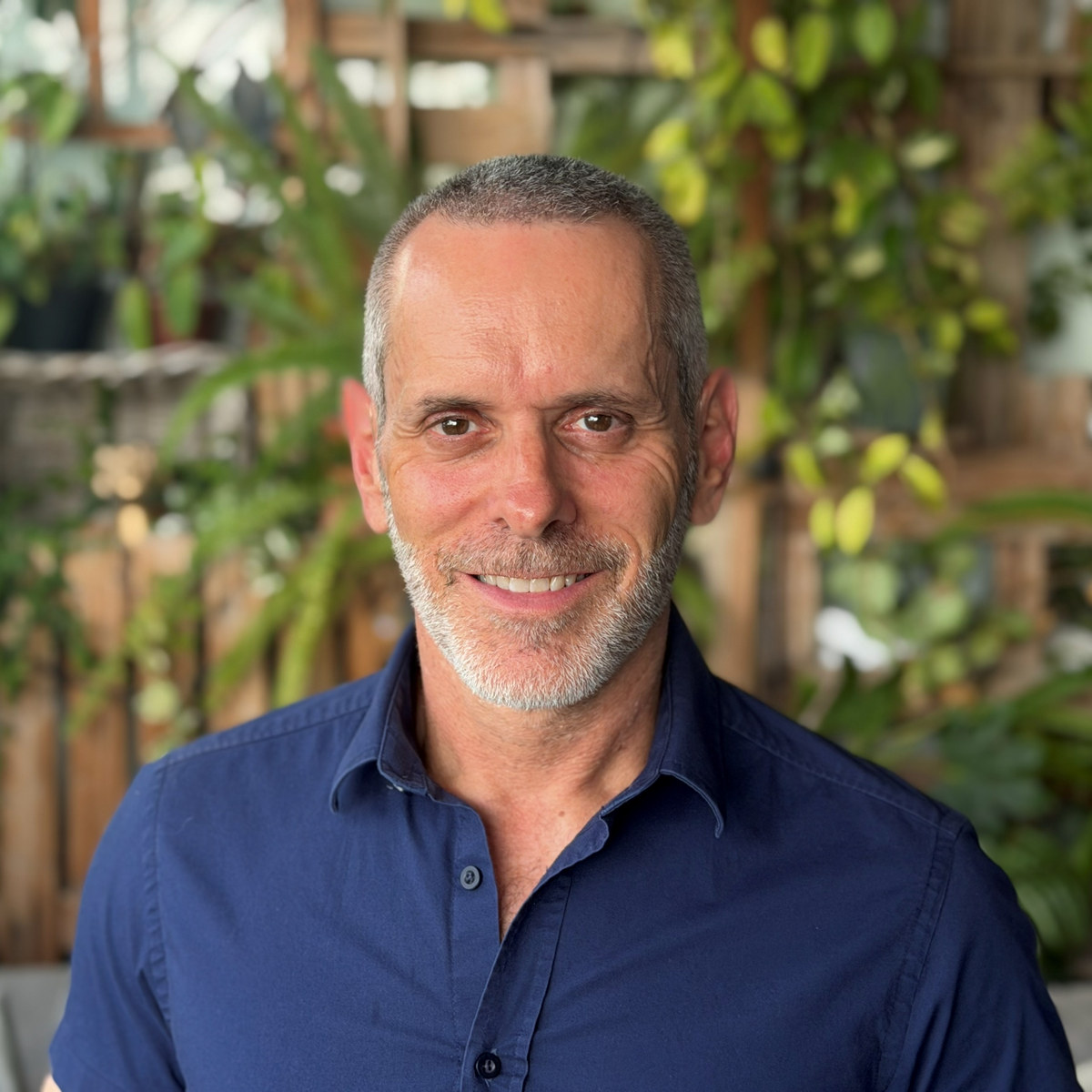 ארז ברוכי מחייך אל המצלמה, בחולצה מכופתרת כחולה כהה, על רקע קיר עץ וצמחייה ירוקה